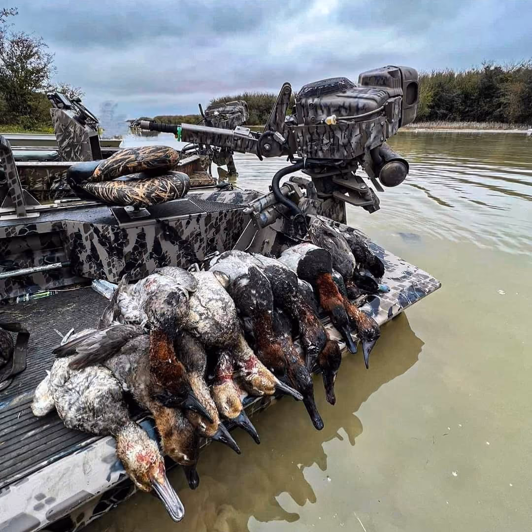 A hunting boat with camouflage patterns holding eight dead ducks lined up on its rear platform near a calm body of water.