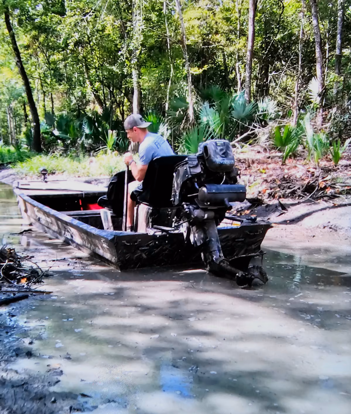 Man wearing a cap sitting in a small motorboat navigating shallow, muddy water through a forested area.