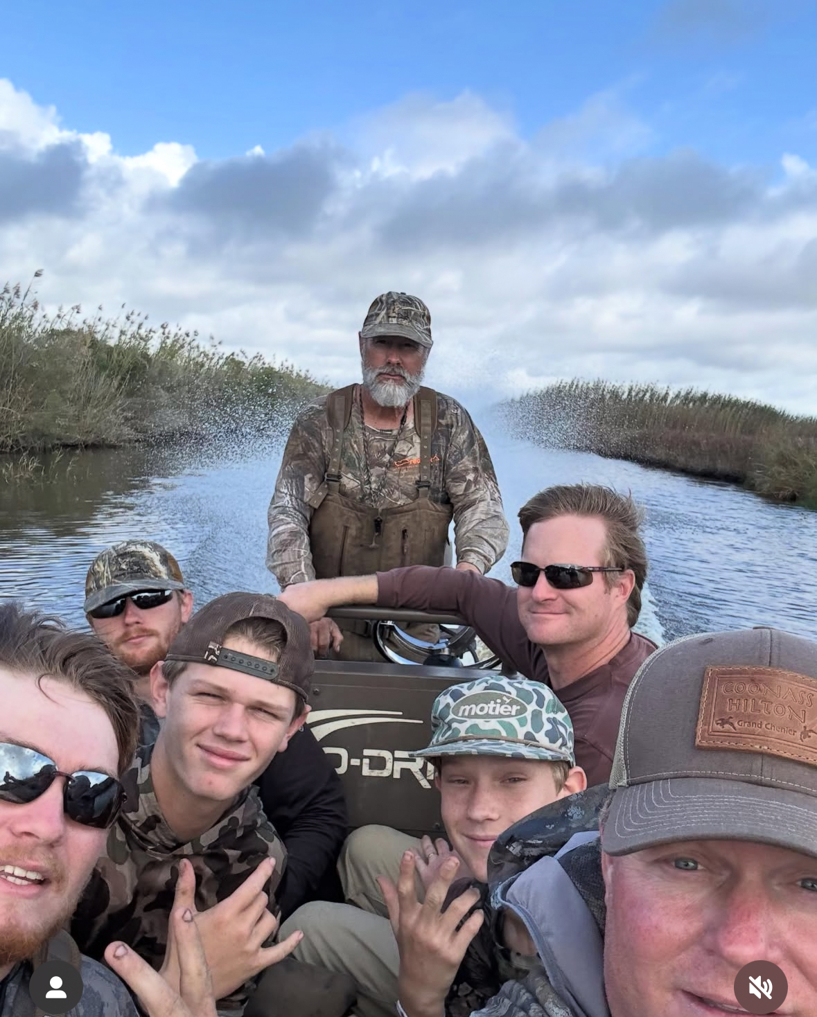 Group of six men wearing camouflage and outdoor clothing riding on a motorboat in a marshy waterway under a partly cloudy sky.