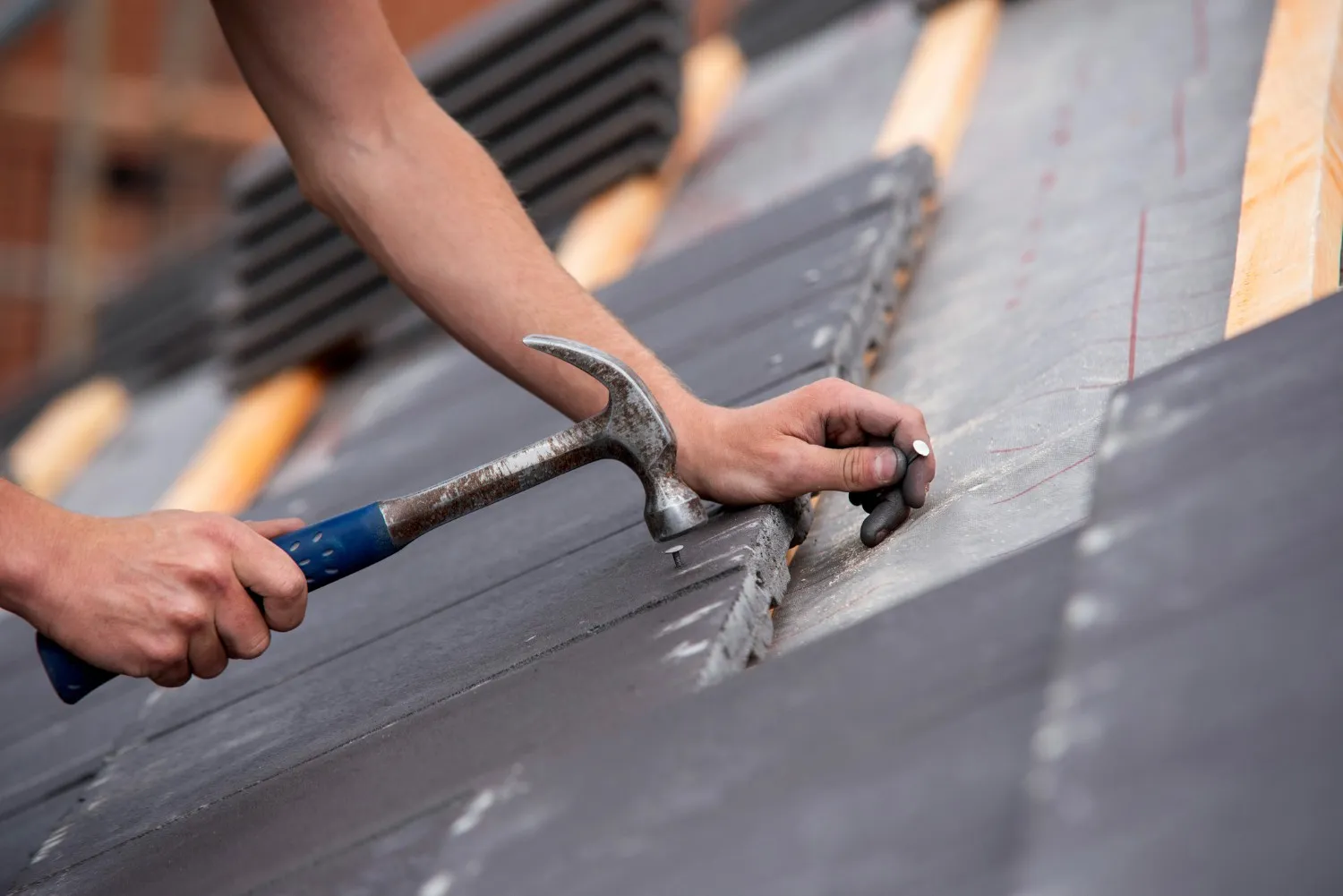 A worker performs roof services by manually hammering a nail into dark grey roof tiles.