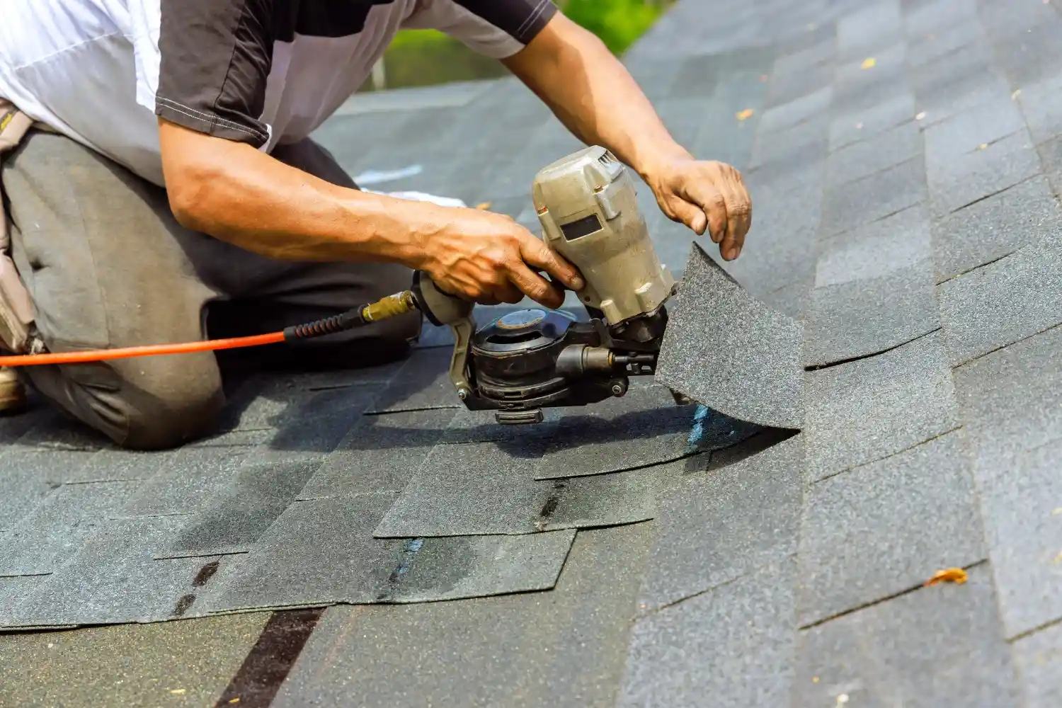A worker performs roof services by using a pneumatic nail gun to install grey asphalt shingles.