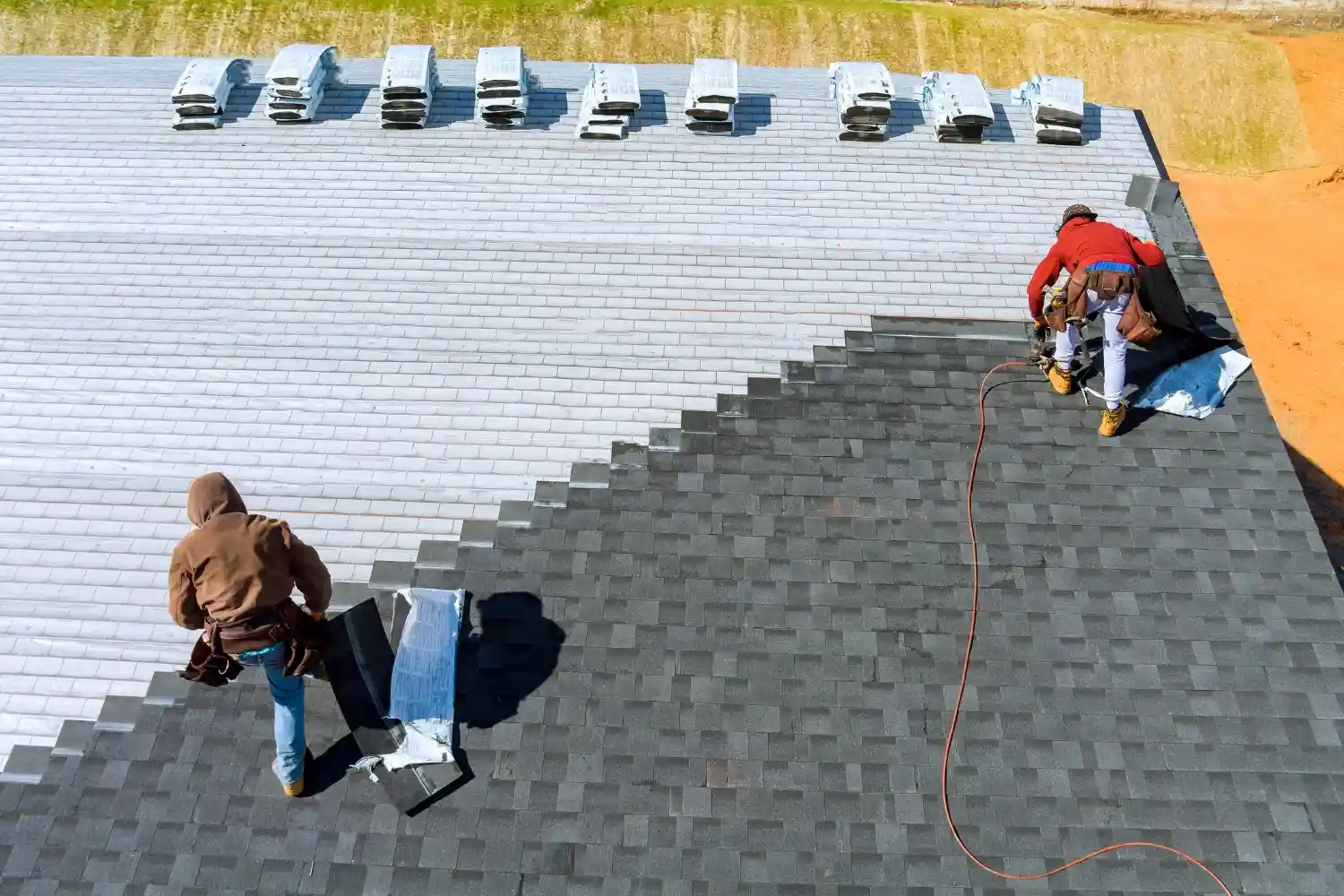 Two workers perform roof services by installing dark asphalt shingles on a large rooftop with stacks of materials nearby.