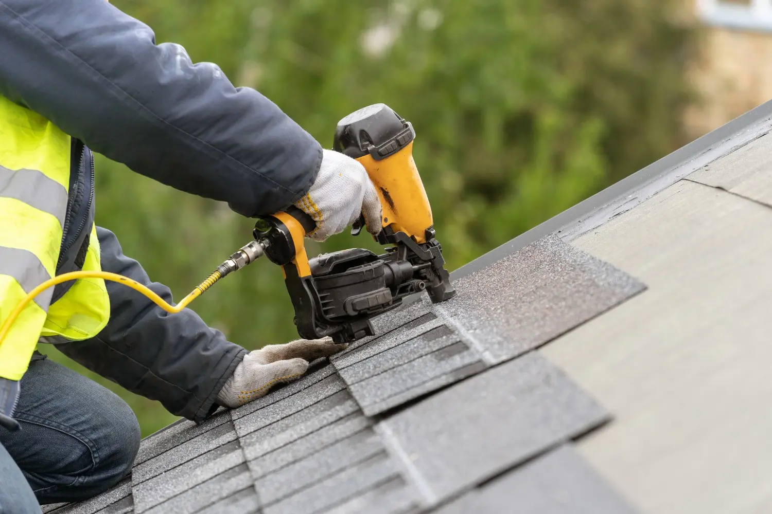 A worker performs roof services by installing shingles using a pneumatic yellow nail gun.