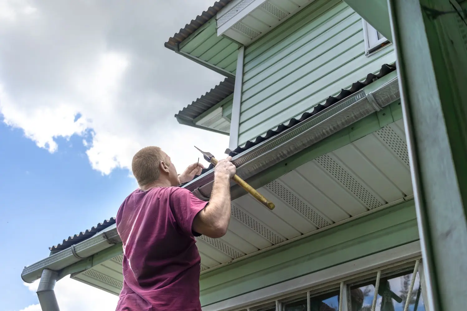 A worker performs gutter services by cleaning the rain gutters of a green residential house using a hand tool.