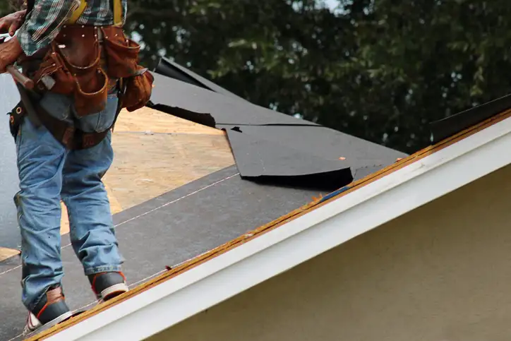 Roofer in tool belt and safety harness lifting dark roofing panel on partially torn-off shingle roof.