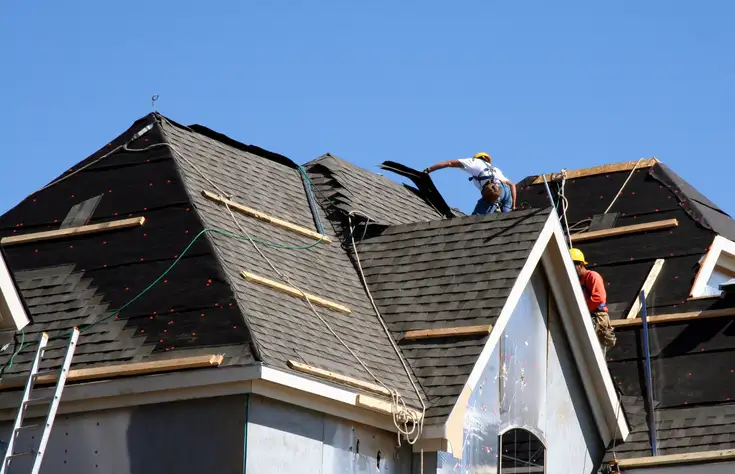 Construction worker in yellow helmet removing old black shingles from a steep residential roof under clear blue sky.
