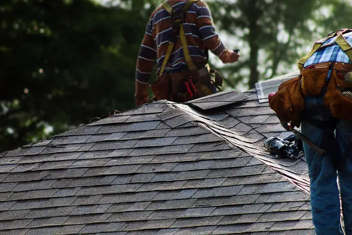 Two roofers wearing tool belts working together to lay overlapping asphalt shingles on steep residential rooftop.