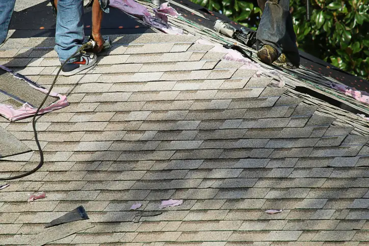 Worker using nail gun to install light gray asphalt shingles on roof, pink underlayment visible underneath.