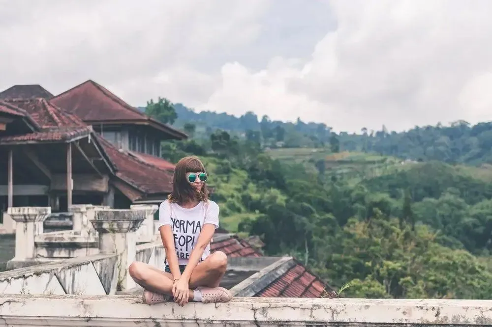 Young woman sitting cross-legged on a rooftop, wearing sunglasses and a t-shirt, with lush green hills and cloudy sky in the background, emphasizing a relaxed outdoor lifestyle.