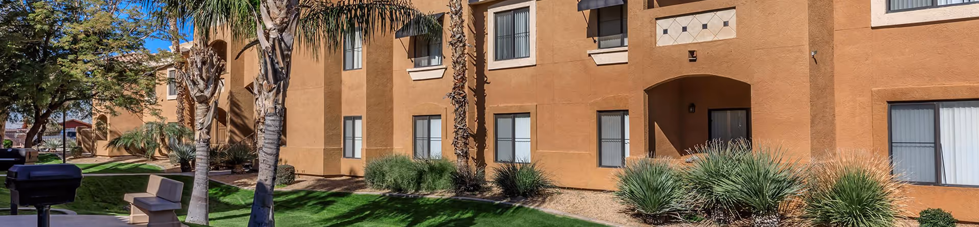 Exterior view of a tan apartment building with multiple windows, palm trees, green grass, and landscaping under a clear blue sky.