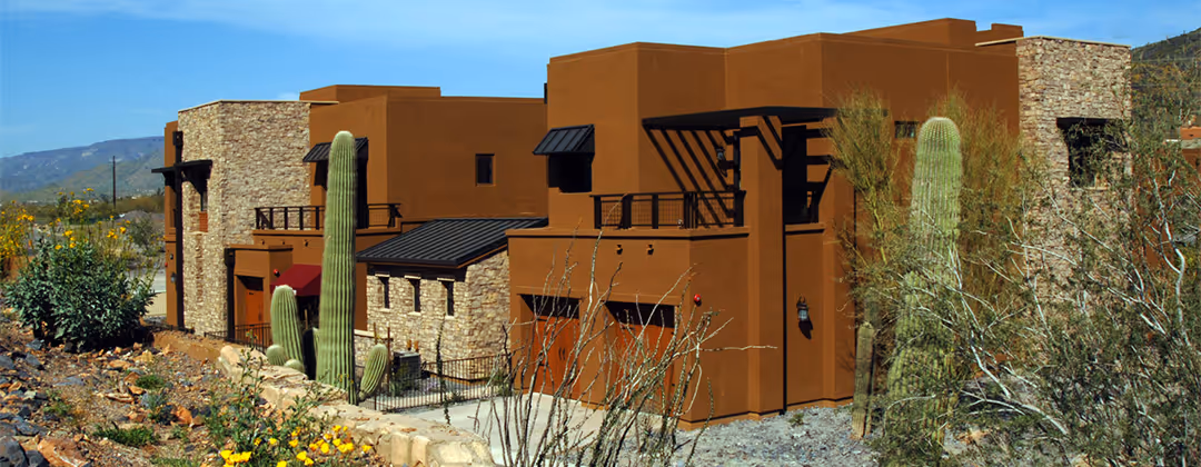Southwestern style brown stucco house with stone accents surrounded by desert plants and tall cacti under a clear blue sky.