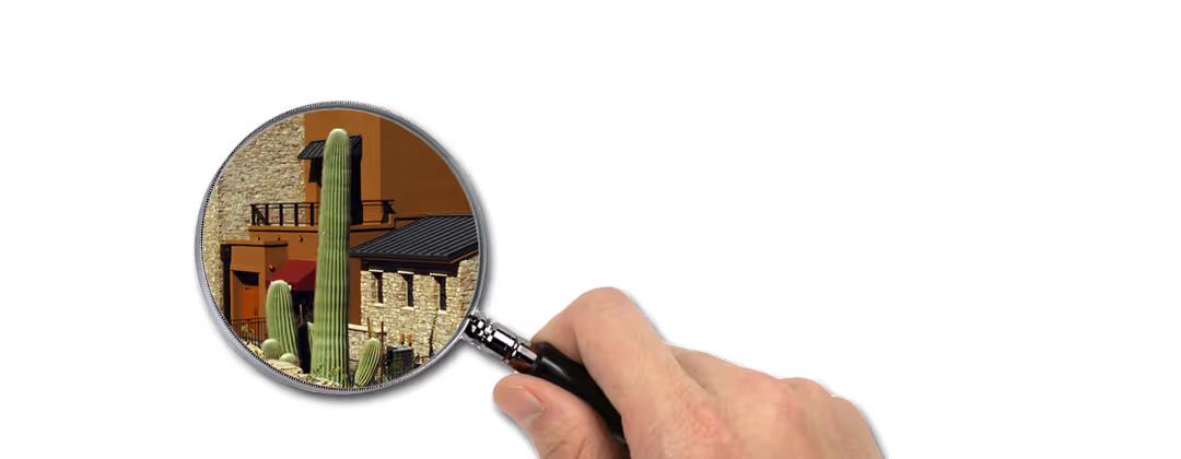 Hand holding a magnifying glass focusing on tall green cacti and buildings with stone and brown walls.