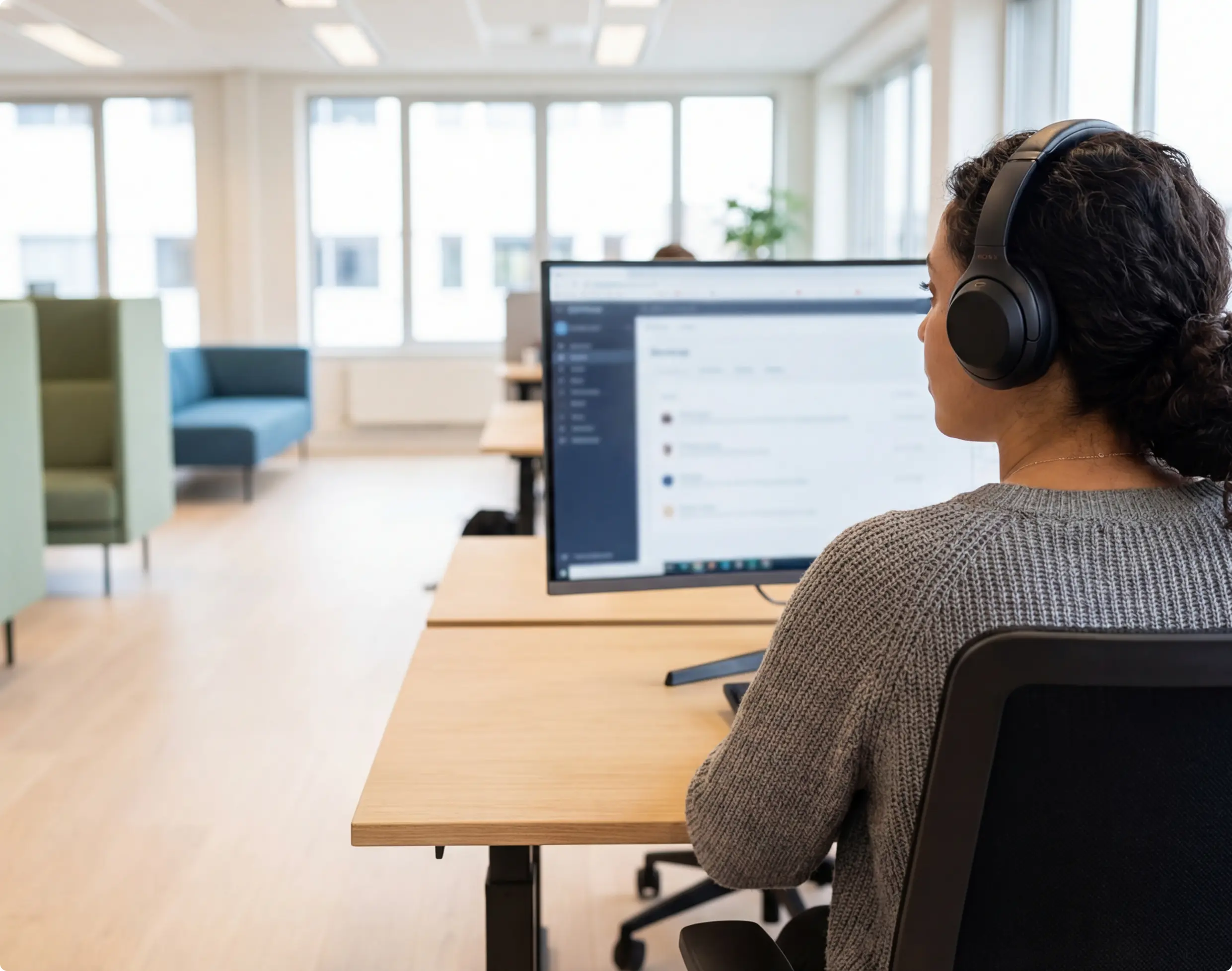 Person wearing headphones and a gray sweater working at a computer in a bright modern office.
