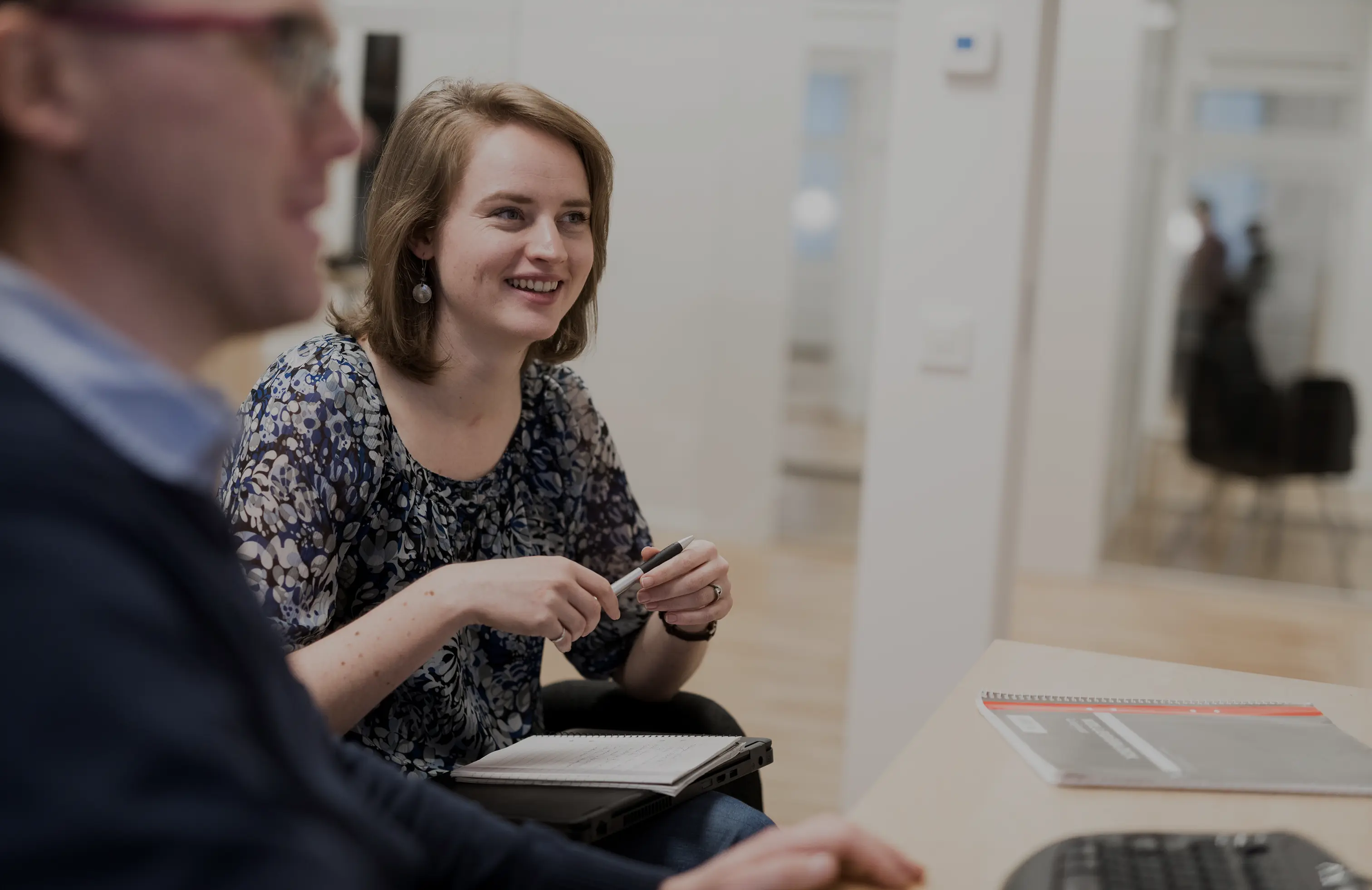 Smiling woman holding a pen and sitting with a notebook on her lap, engaged in conversation in an office setting.