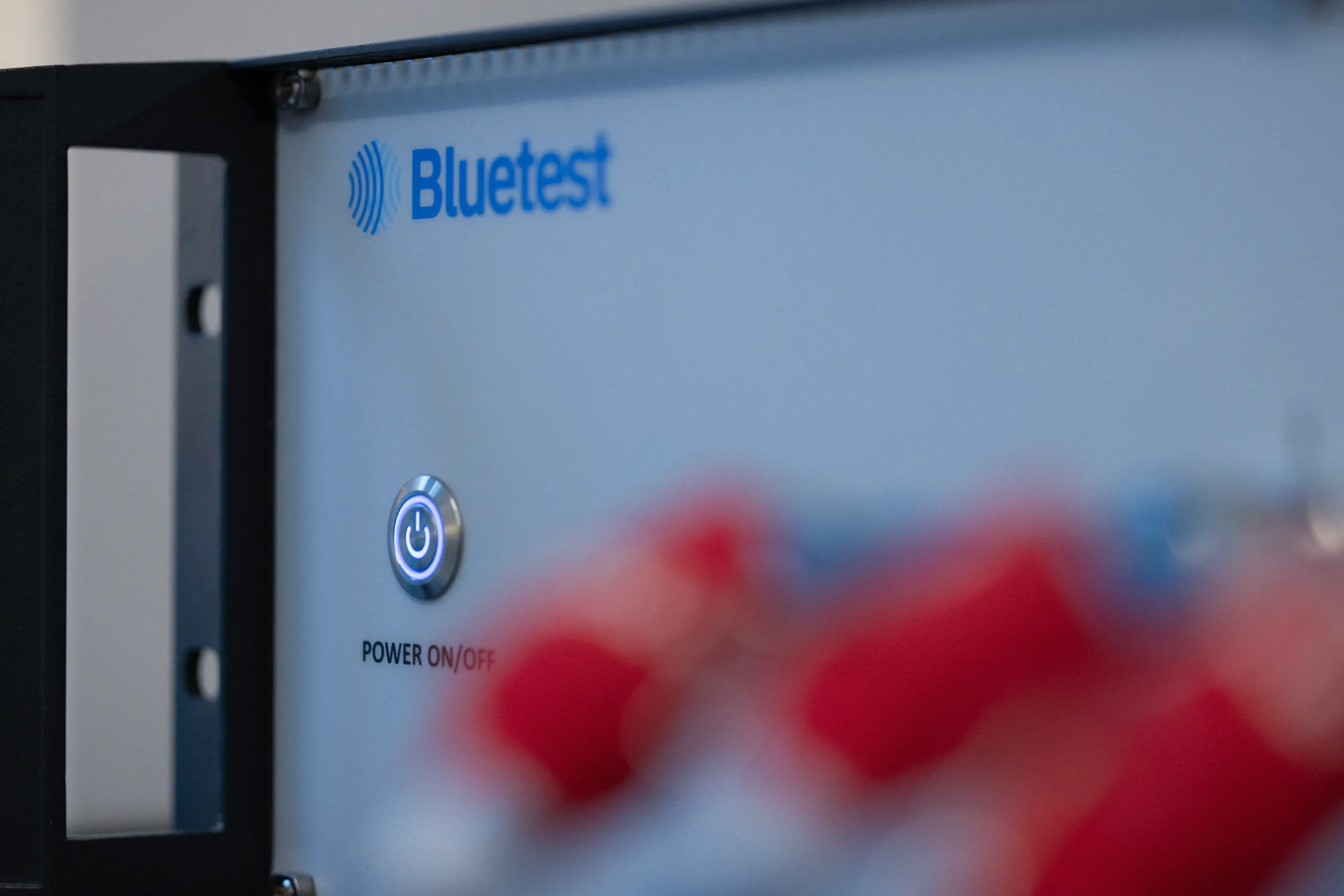 Close-up of a Bluetest device power button glowing blue with blurred red connectors in the foreground.