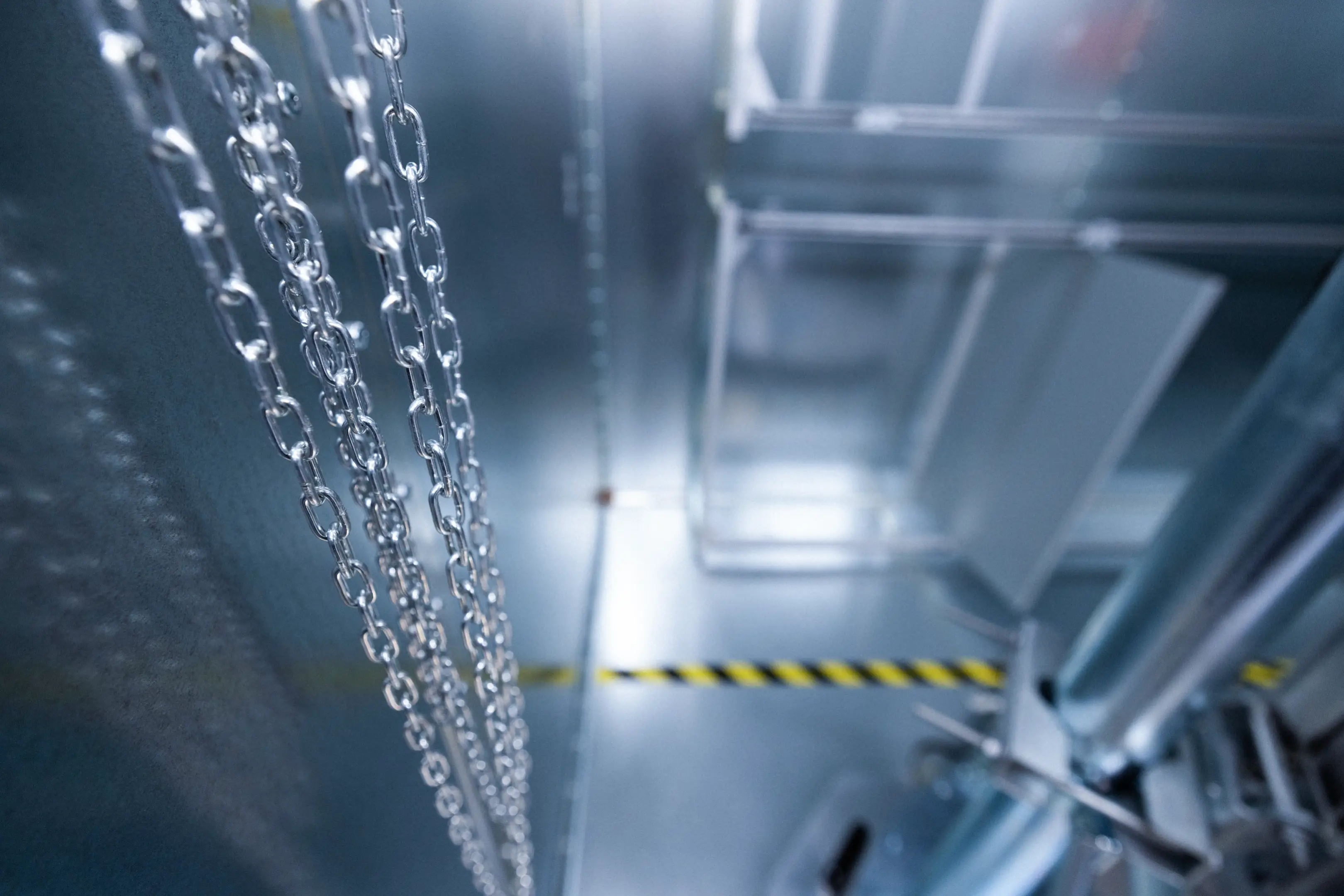 Close-up of metal chains hanging in an industrial setting with blurred machinery and a yellow-black caution tape in the background.