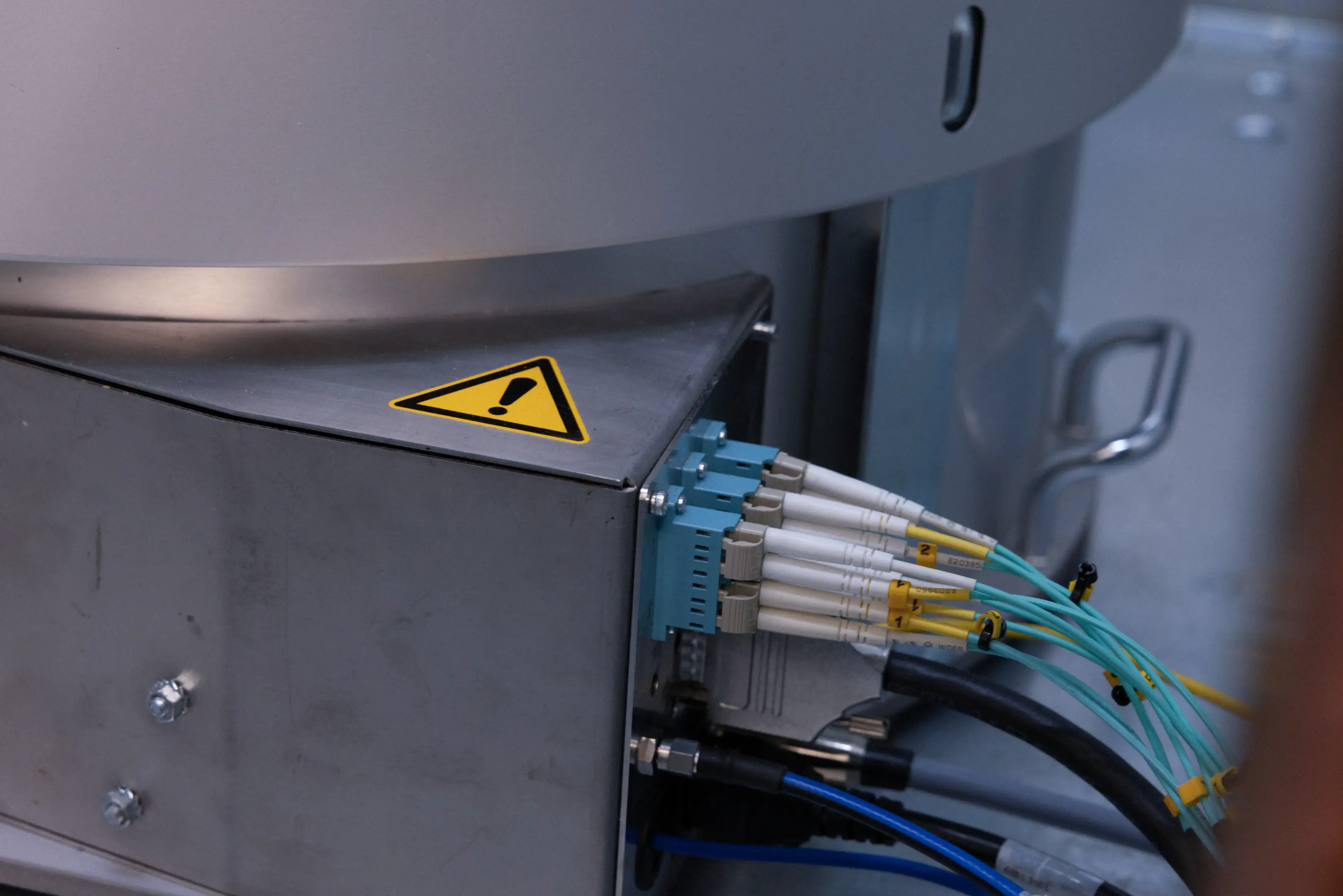 Close-up of a metal electrical box with fiber optic cables connected, featuring a yellow warning triangle.