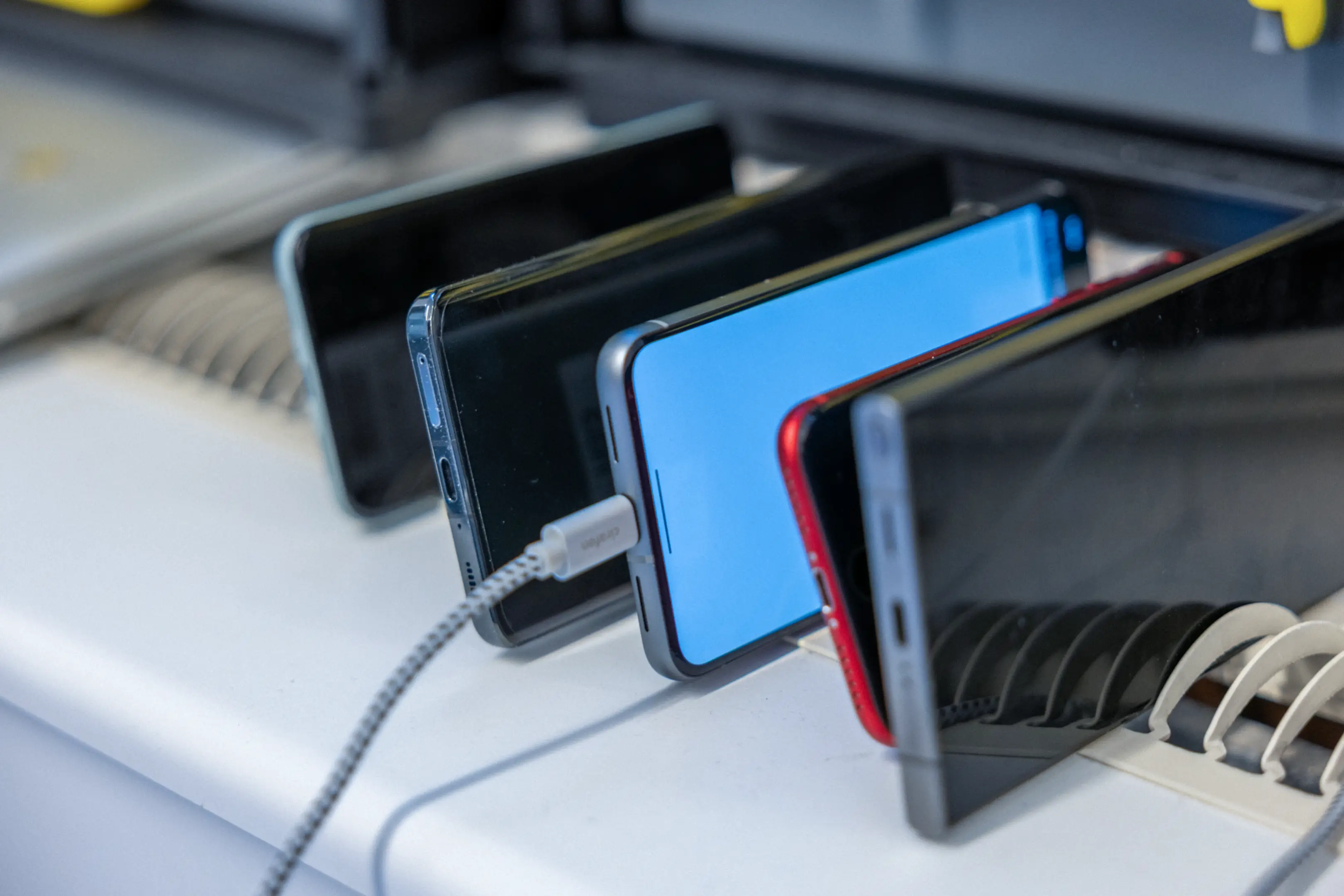 Multiple smartphones standing vertically in a row on a white surface, with one phone plugged into a charging cable.