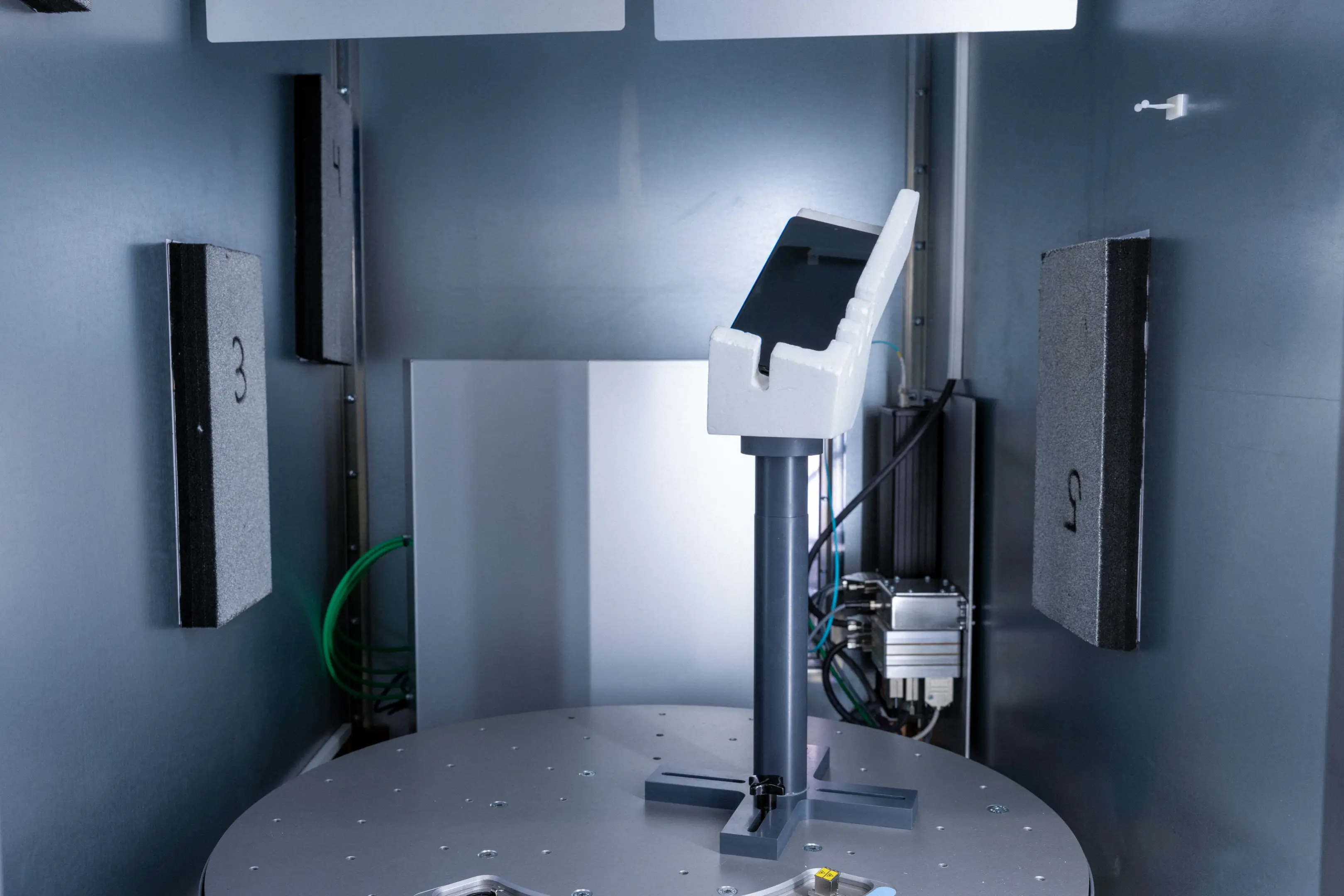 Laboratory testing device holding a smartphone in a white foam grip inside a metal chamber with numbered foam pads on the walls.