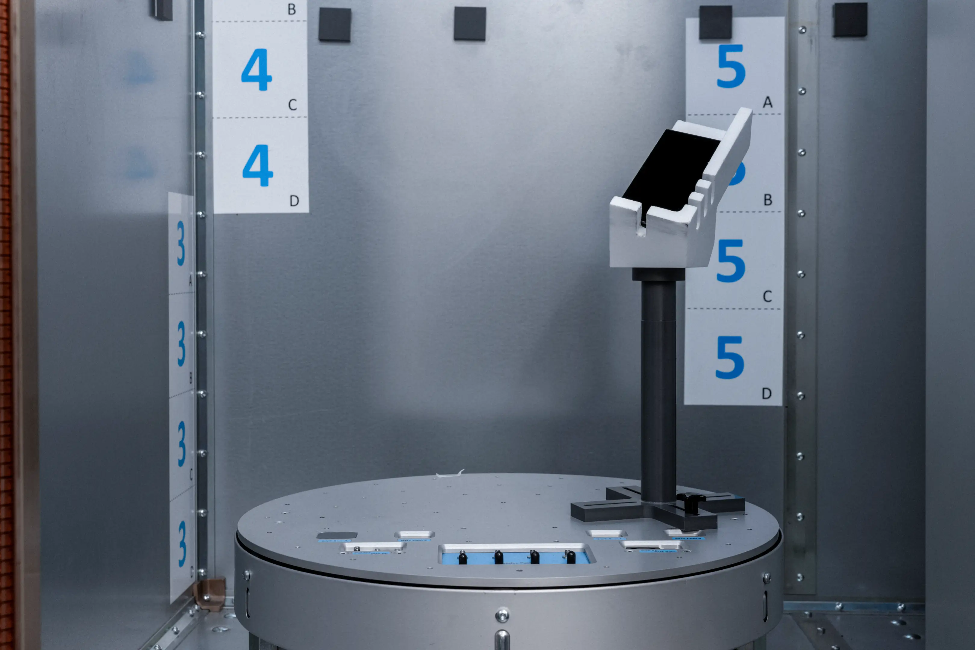 Laboratory setup with a gray circular metal platform and a black and white device mounted on a stand inside a metallic enclosure with numbered measurement scales.