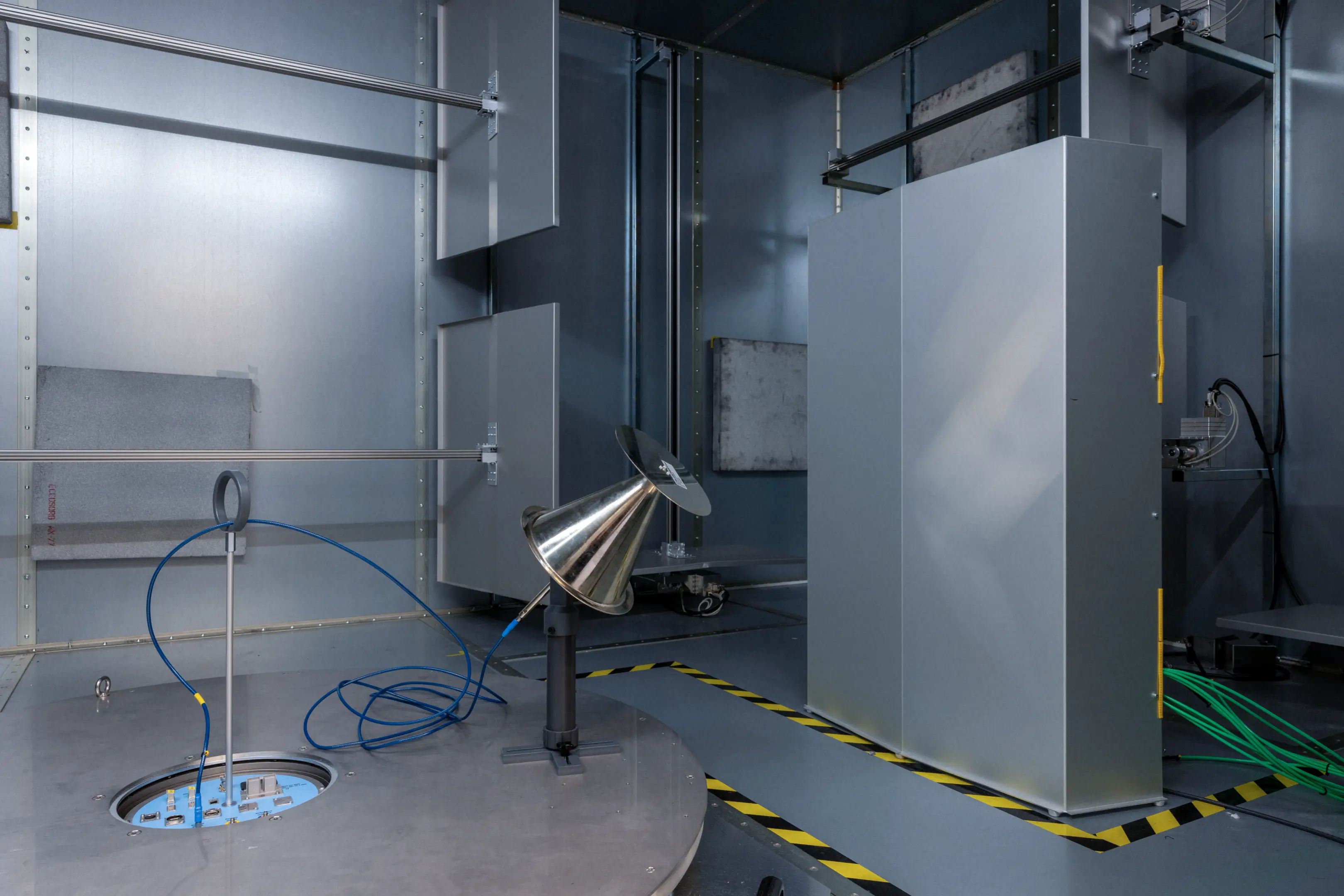 Interior of an anechoic chamber with a metal rotating platform, a conical antenna, and gray acoustic panels on the walls.
