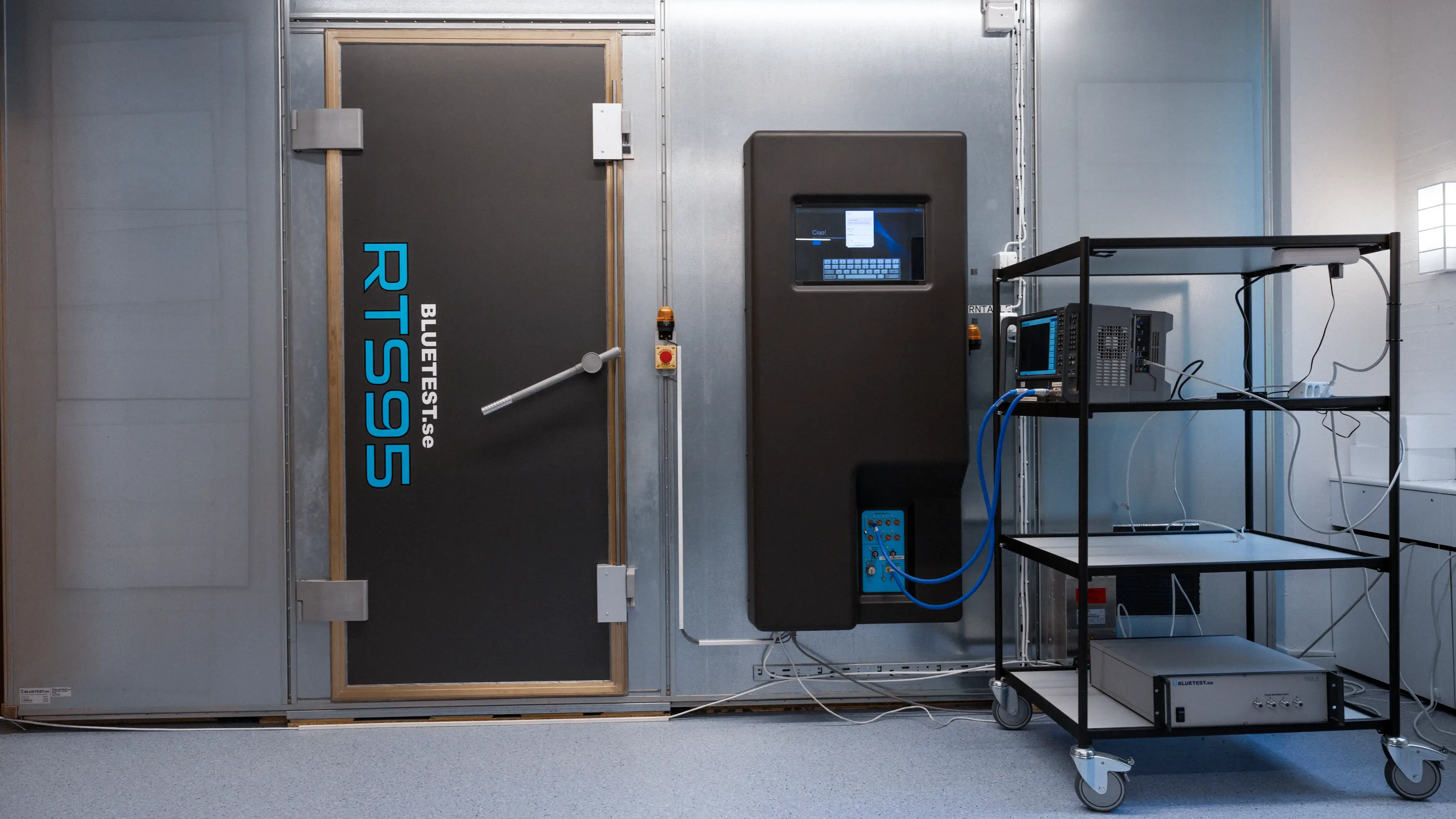 Industrial testing room with a black door labeled RTS95 and electrical testing equipment on a mobile metal cart.