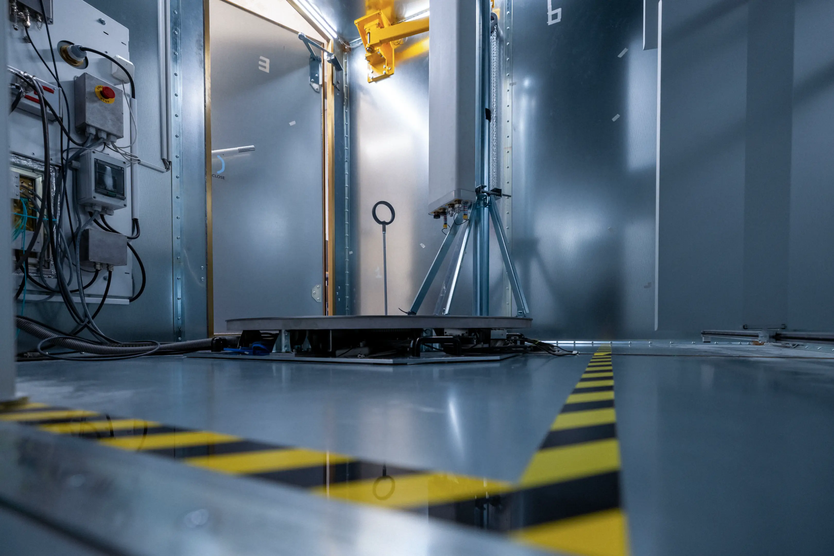 Interior view of industrial testing chamber with metal walls, control panel, and a platform with a tripod-mounted device.
