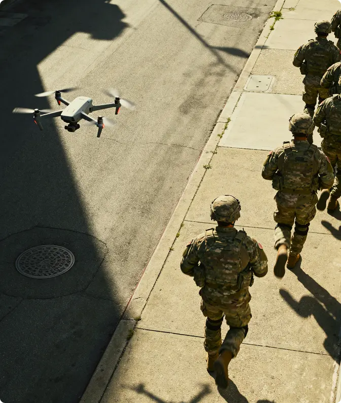 A drone flying above a street next to soldiers in camouflage uniforms walking on a sidewalk.
