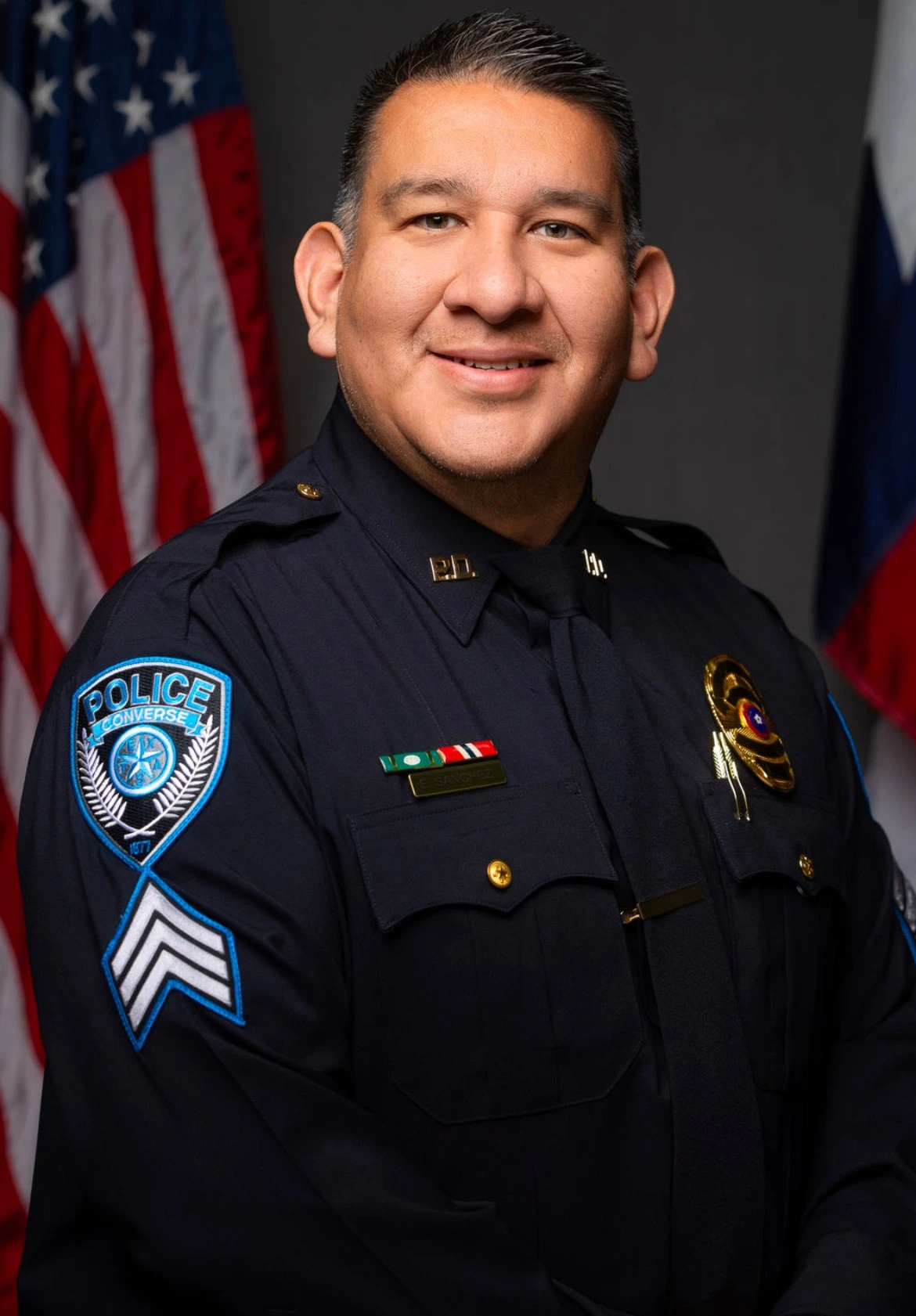 Smiling police officer in dark uniform with Converse Police patch and rank chevrons, posing with American flag in background.