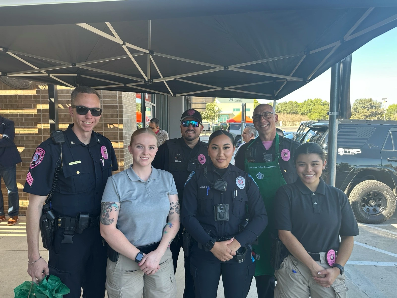 Group of six police officers and staff posing under a tent outdoors, smiling at the camera.