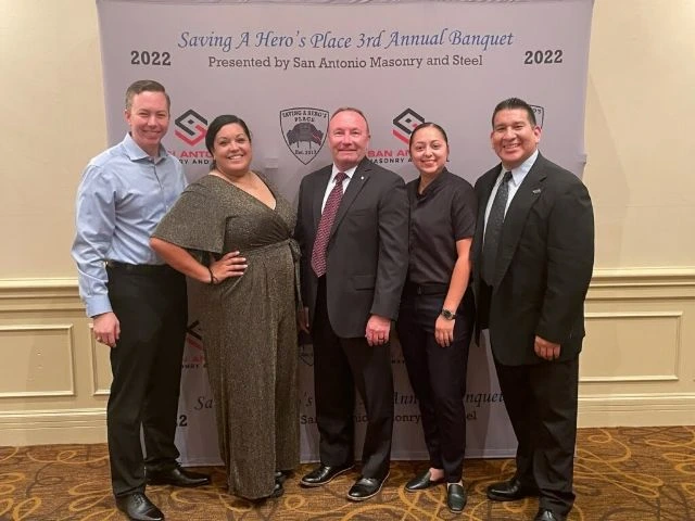 Five people dressed in formal attire posing in front of a backdrop for the Saving A Hero's Place 3rd Annual Banquet presented by San Antonio Masonry and Steel in 2022.