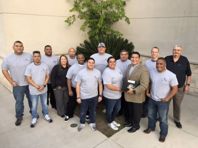 Group of twelve adults standing outdoors on a concrete surface near a green plant against a beige wall, most wearing matching light gray t-shirts with a logo.