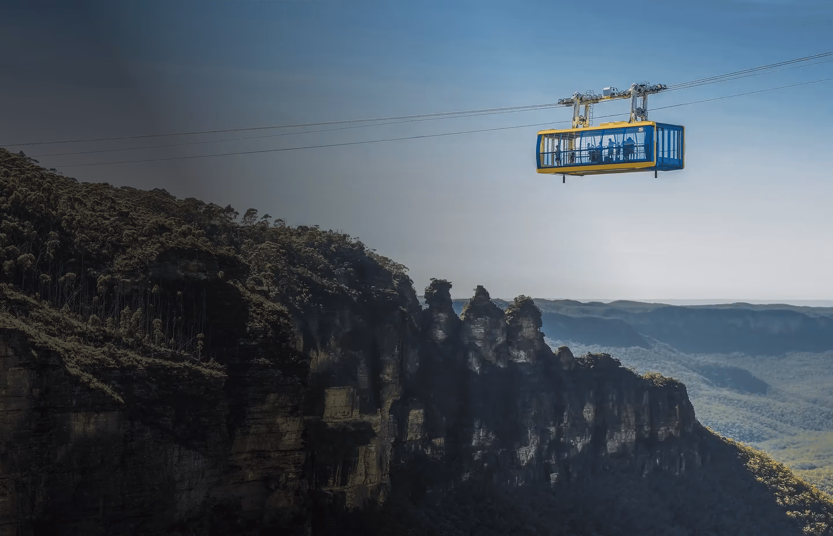 Cable car with passengers traveling over a forested mountain cliff with a clear sky background.