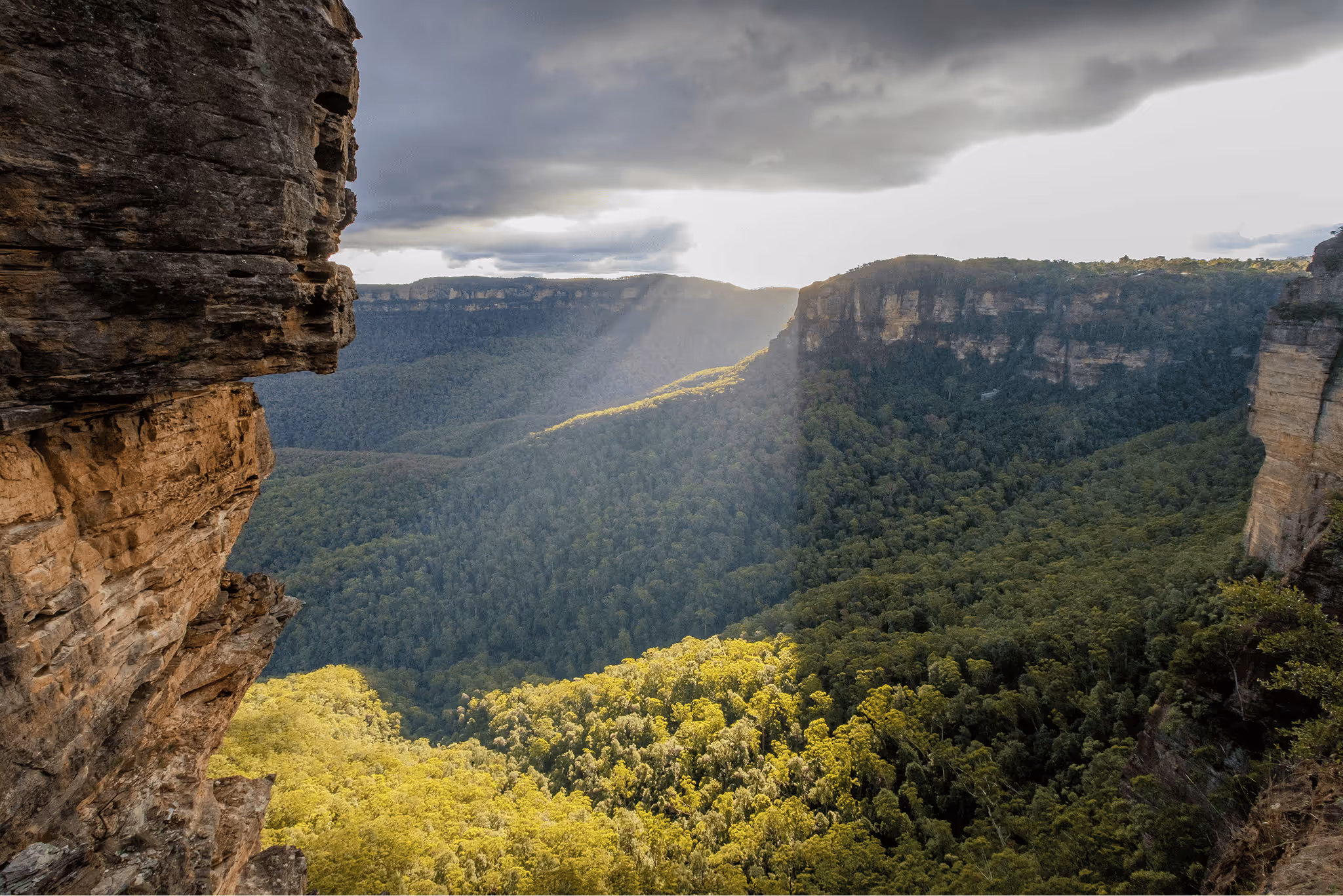Sunlight beams through clouds over a dense green forest valley surrounded by steep rocky cliffs.
