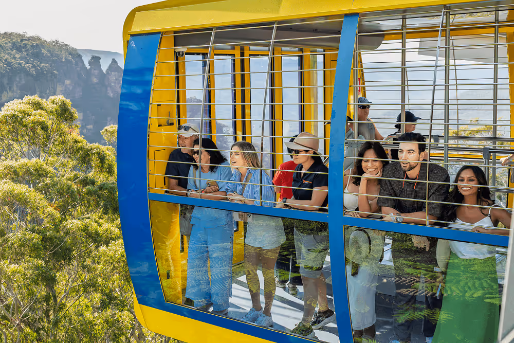 Group of people enjoying a scenic view from a yellow and blue cable car above green forest with mountains in the distance.