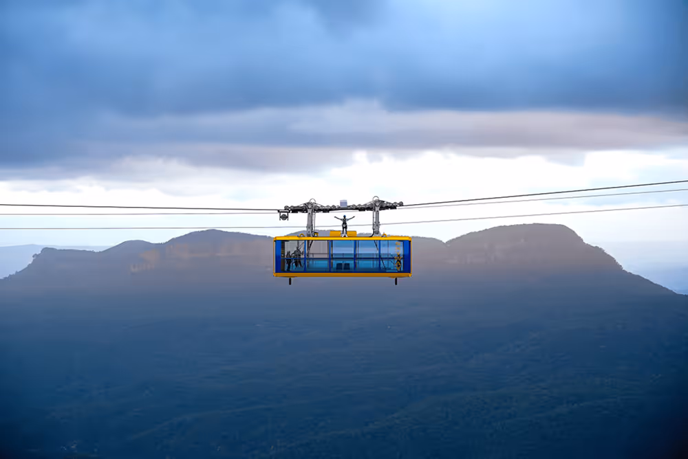 Yellow and blue rectangular cable car suspended on cables over a mountainous landscape under a cloudy sky.