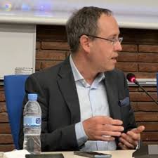 Man in a suit and glasses speaking at a table with a microphone, water bottle, and phone in front of him.