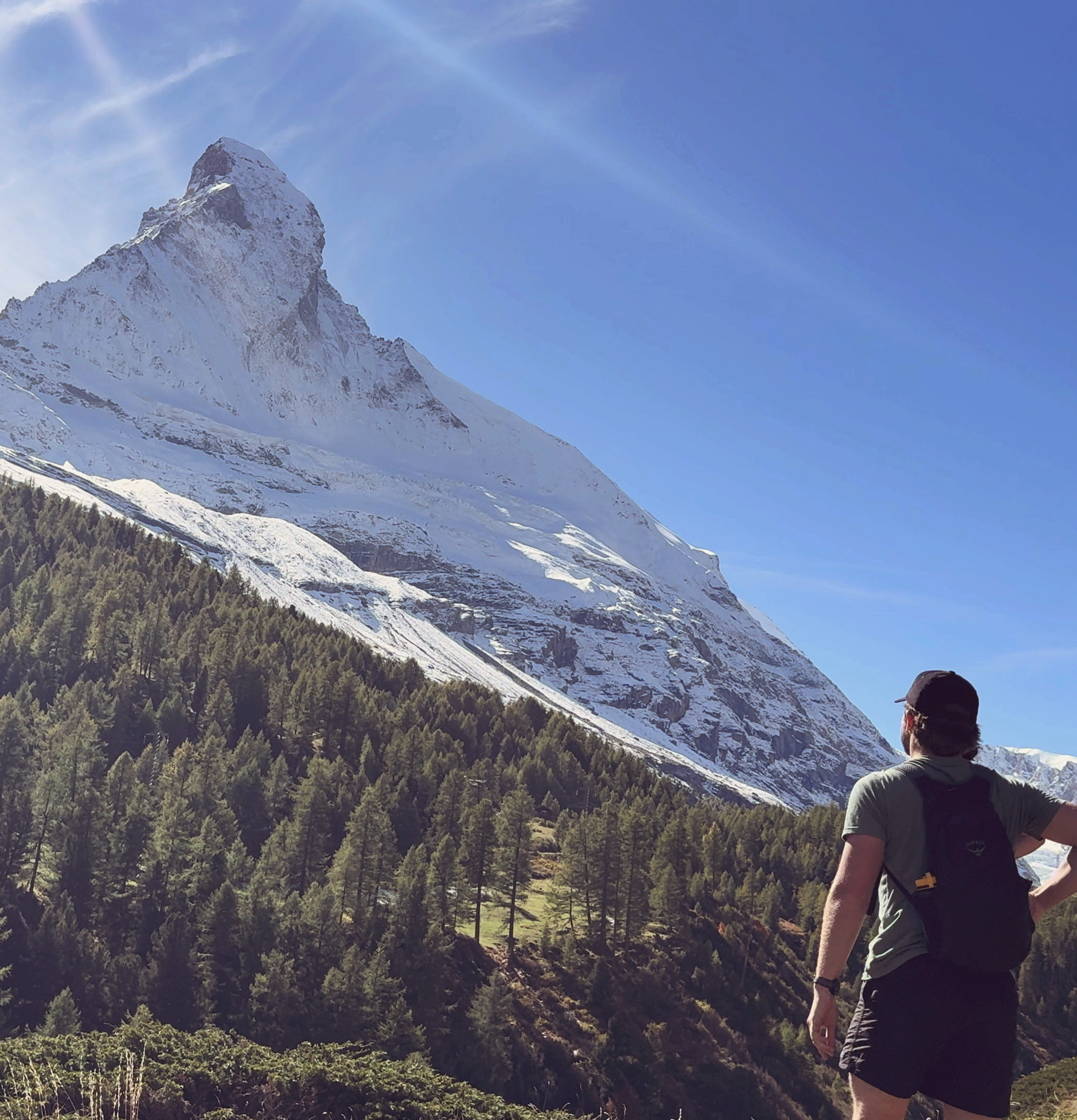 Hiker standing on a mountain trail overlooking a forested slope and a large snow-covered peak under a clear blue sky.