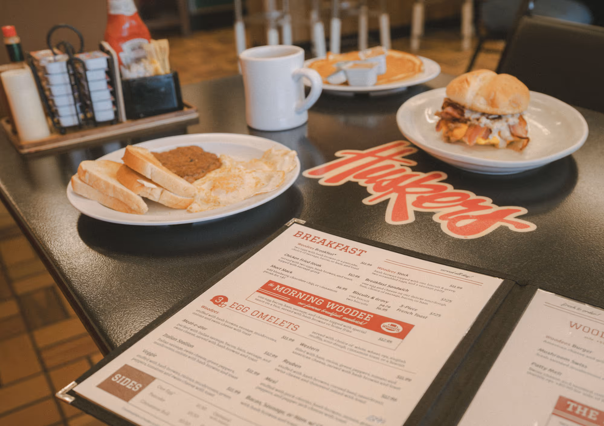 Breakfast dishes with eggs, toast, sandwich, coffee cup, and pancakes on a table with a Woodee's menu.