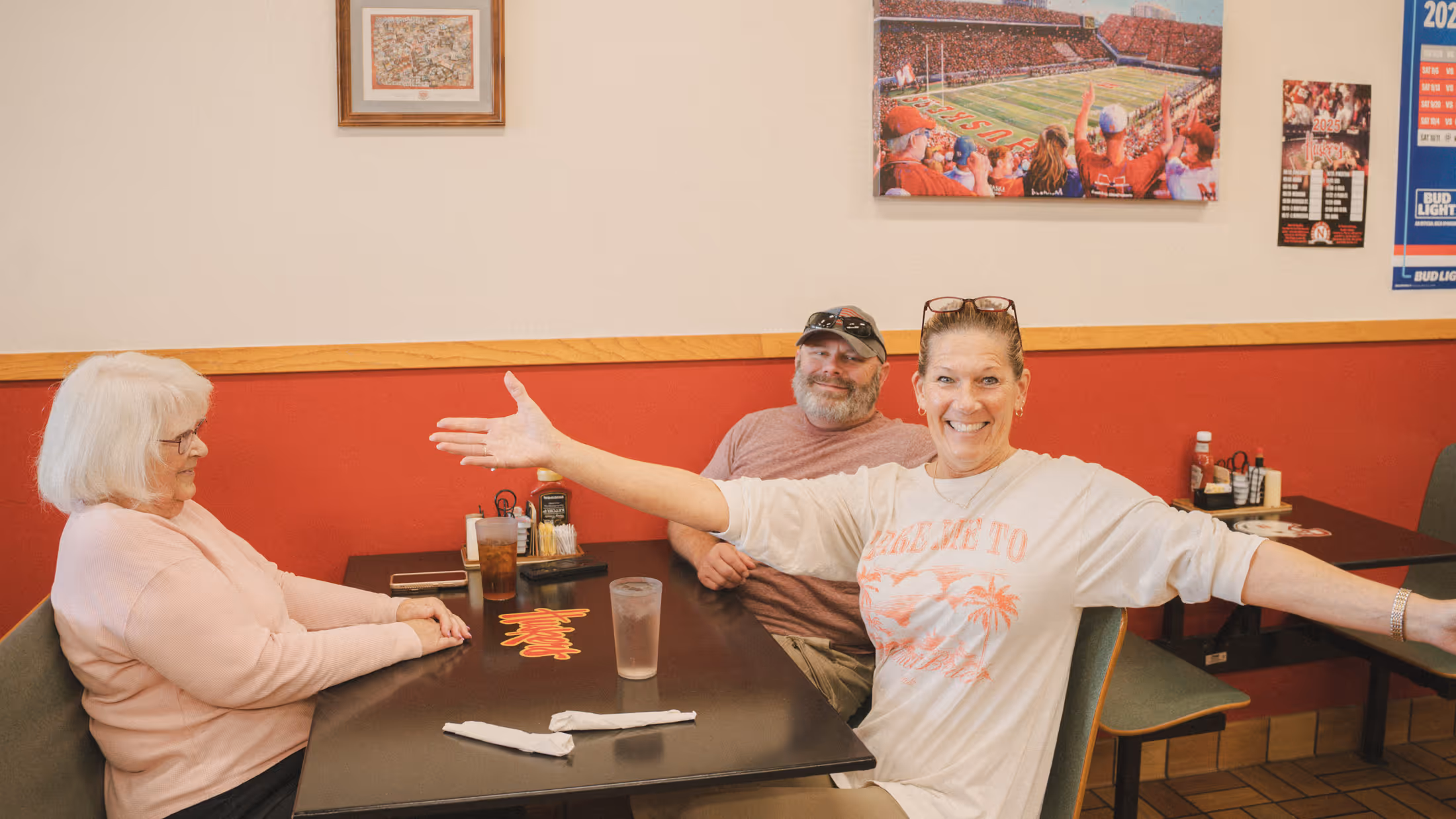 Lynette and customers sit at a table with red walls and sports posters in the background.