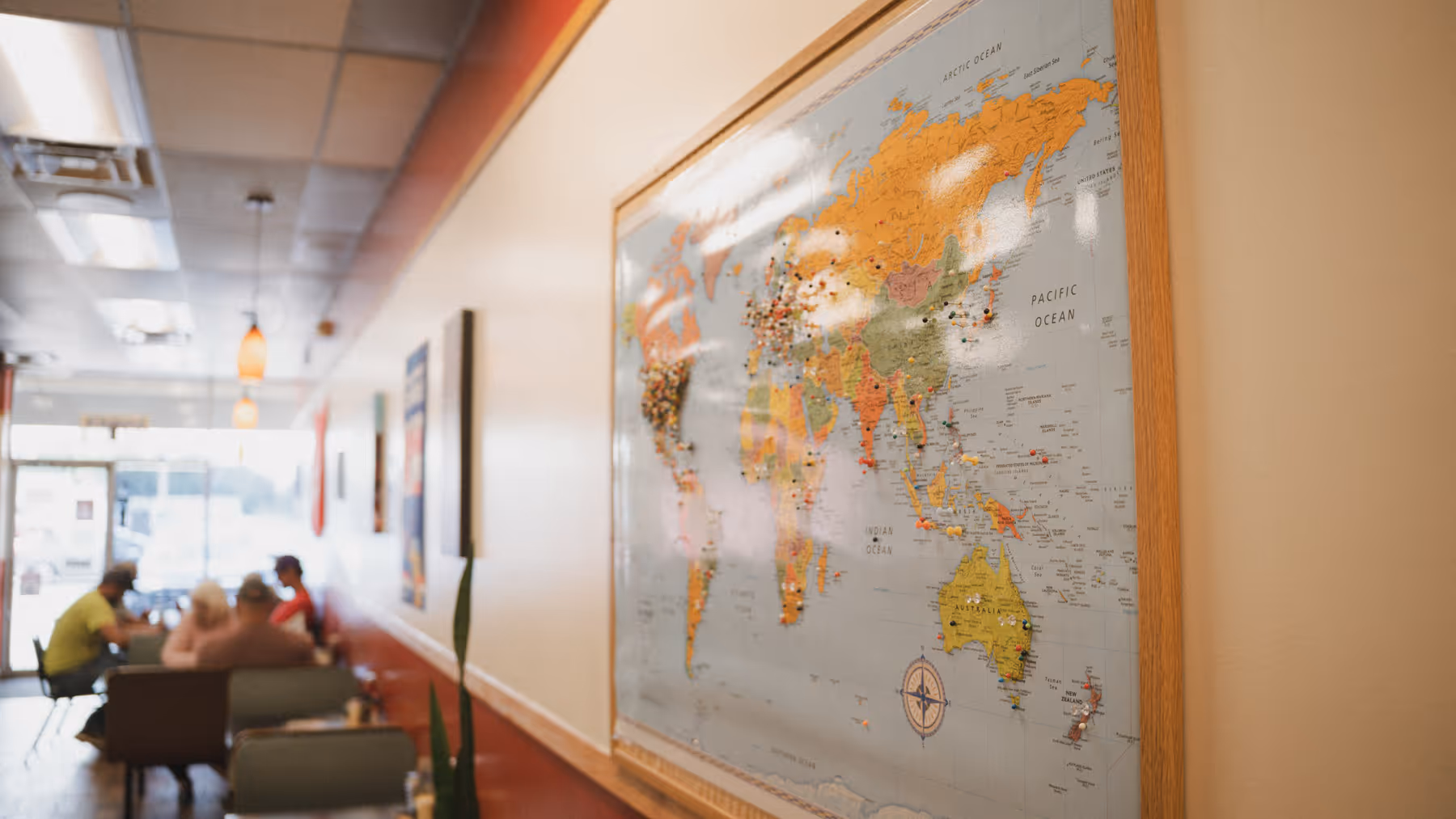 World map with colored pins on wall in a cafe where people sit at tables in the background.