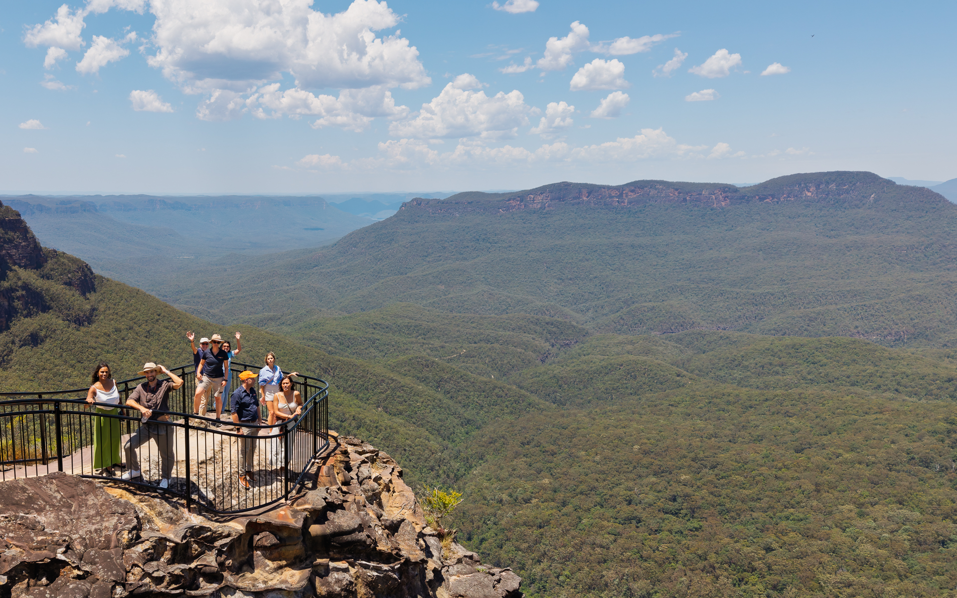 A group of people enjoying the breathtaking panoramic view from a lookout point atop a rocky cliff, overlooking a vast expanse of lush green forest and distant mountain ranges under a bright blue sky with scattered clouds.