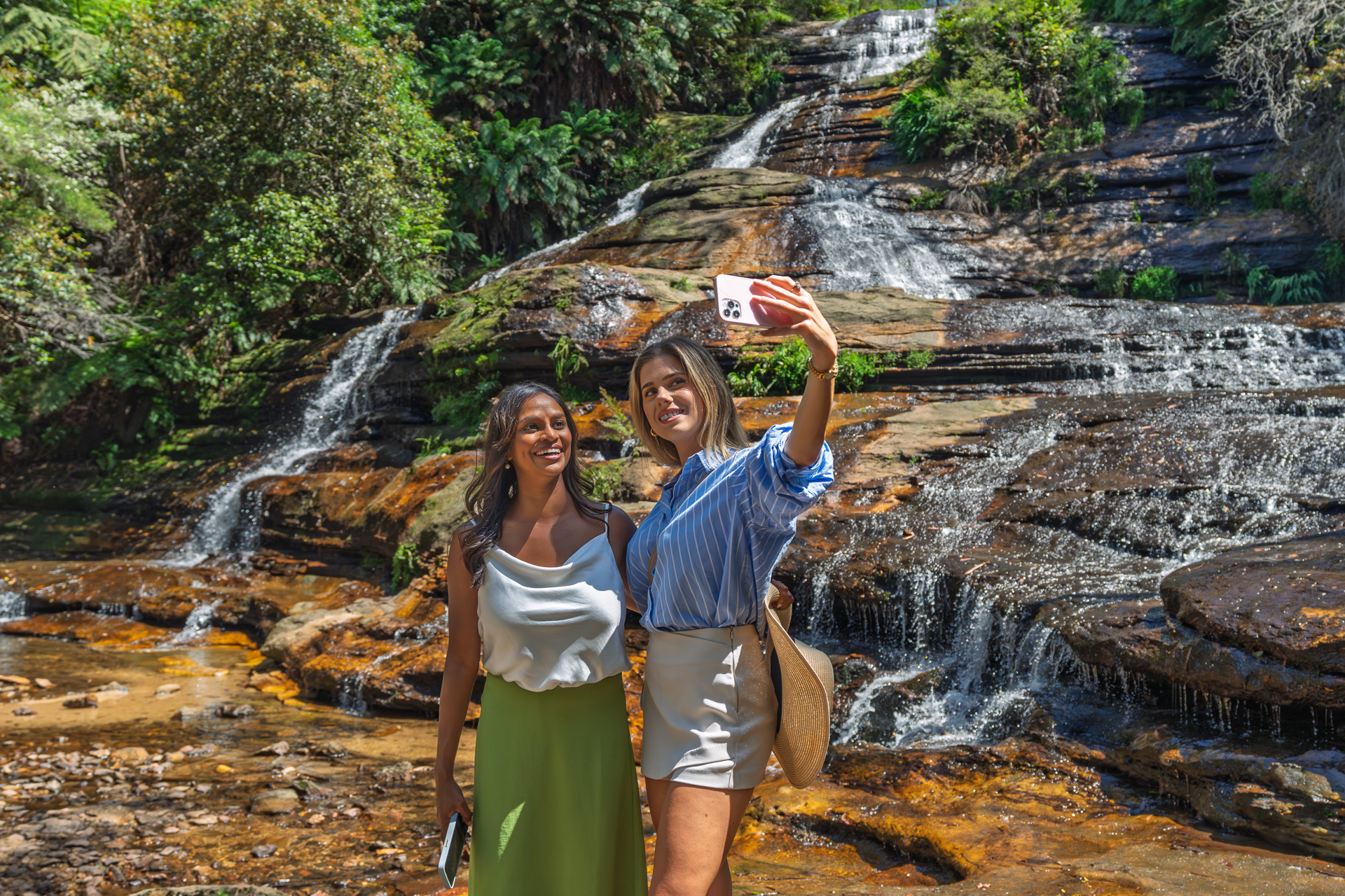 Two women stand in front of a rocky waterfall, smiling and taking a selfie together on a sunny day surrounded by greenery.