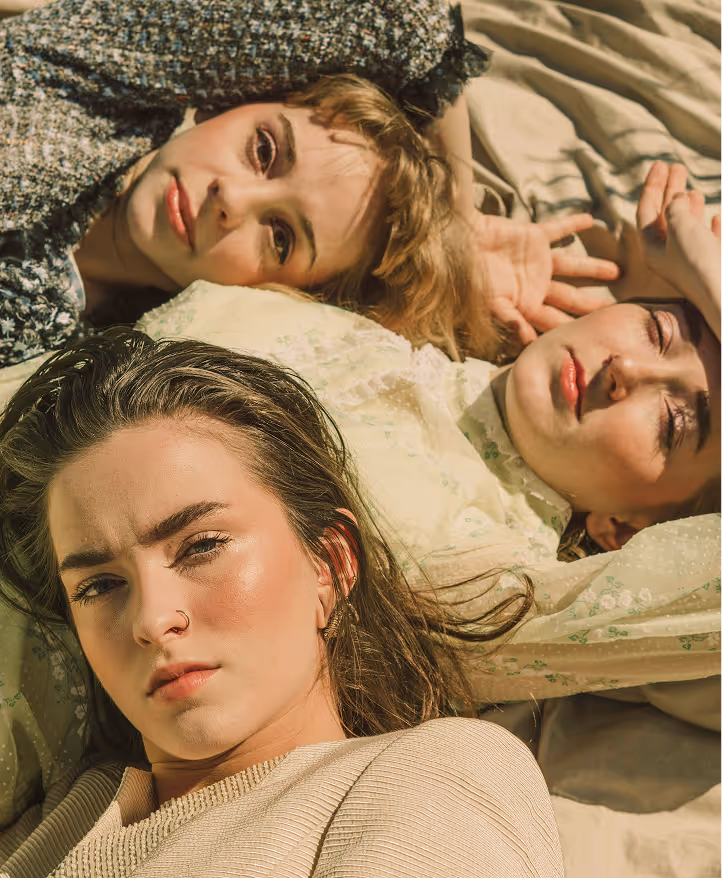 Three women lying down close together in sunlight, with relaxed and thoughtful expressions.