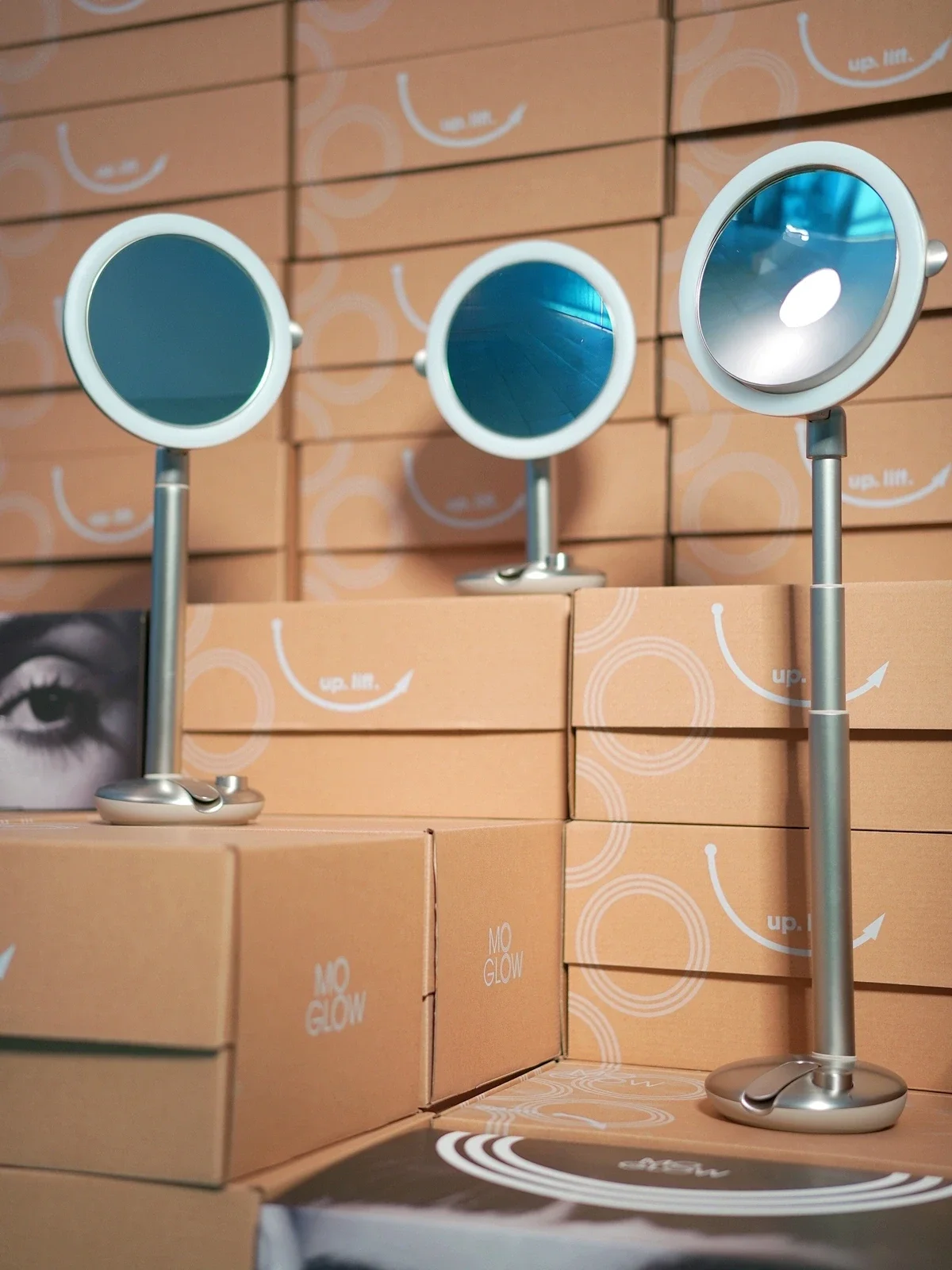 Three round MoGlow mirrors with silver stands placed among stacked cardboard boxes labeled.