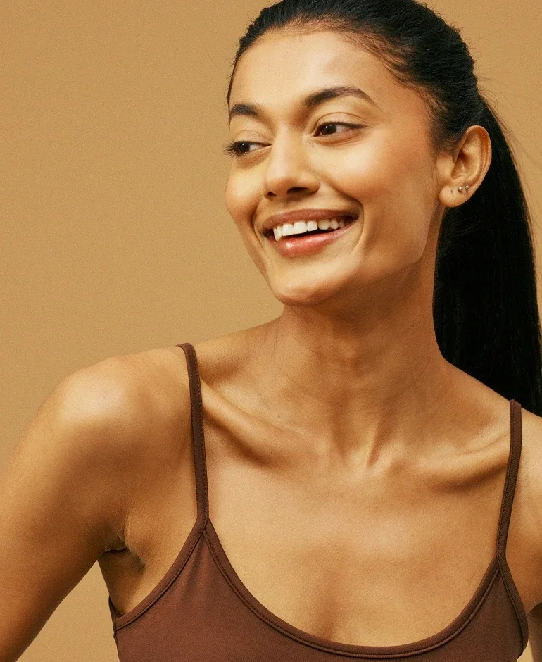 Smiling woman with dark hair in a brown spaghetti strap top against a beige background.