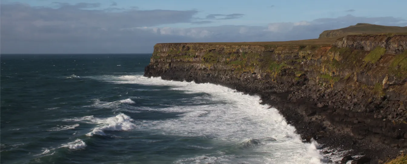Cliffs covered with patches of grass and moss beside a rough ocean with waves crashing against the rocky shore under a partly cloudy sky.