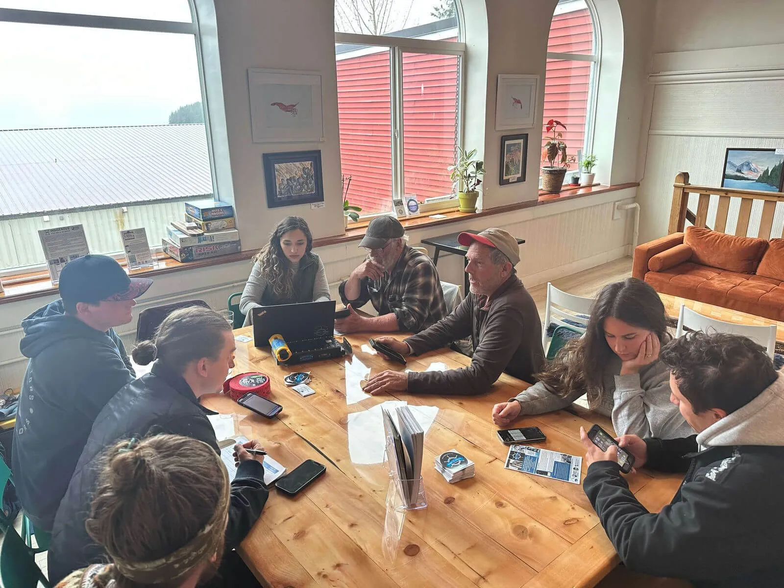 Seven people sitting around a wooden table in a well-lit room with large arched windows, engaged with laptops, smartphones, and notes, with board games stacked on the windowsill and artworks on the walls.