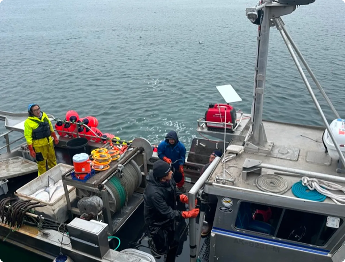 Four people wearing waterproof gear working on a boat equipped with fishing and marine equipment, with water visible in the background.