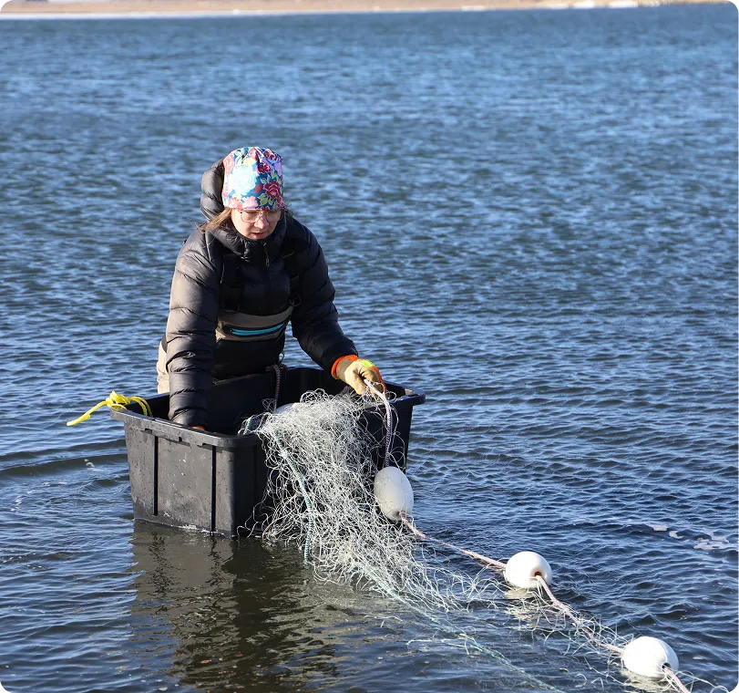 Person wearing a floral hat, glasses, and a black jacket sitting in a small black container floating in water, handling a fishing net with white floats.
